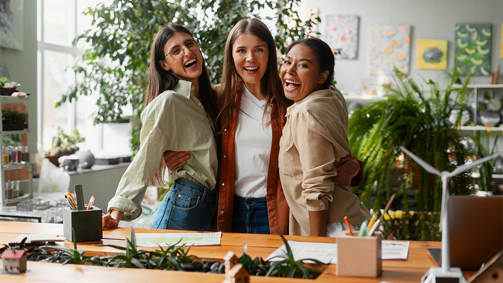 Três mulheres empreendedores abraçadas e felizes em escritório representando a relação entre empreendedorismo e empoderamento feminino