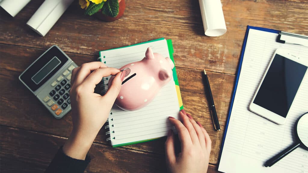 Mão de mulher colocando moeda em cofre em formato de porco, representando a ideia de reserva financeira
