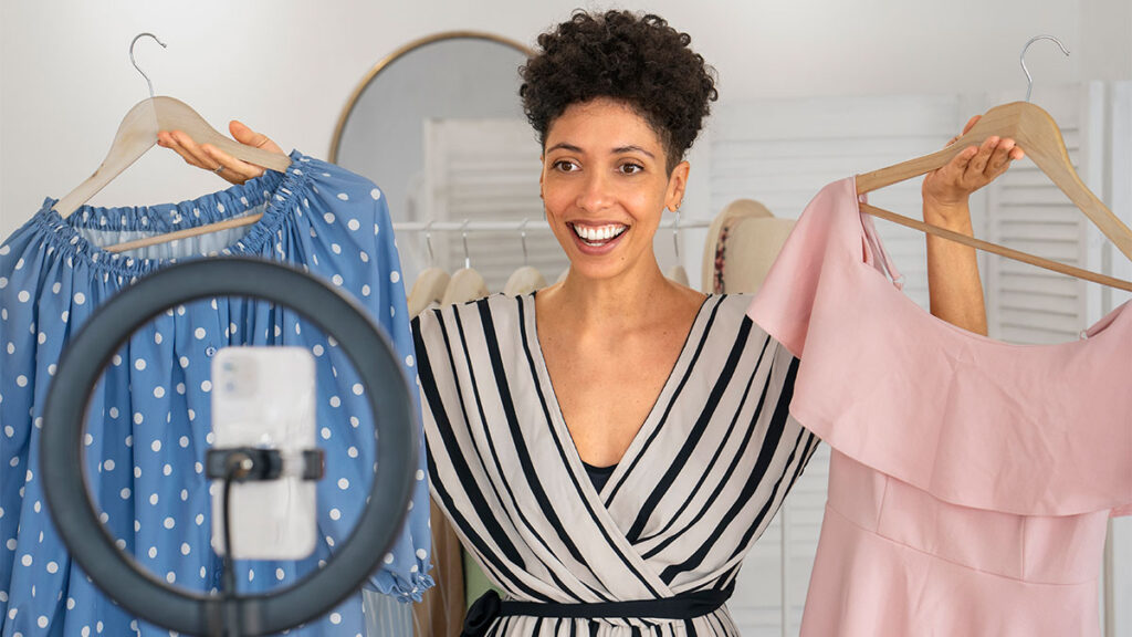 Mulher sorridente grava vídeo mostrando dois vestidos, ilustrando dicas de como criar uma segunda renda com vendas online de roupas.