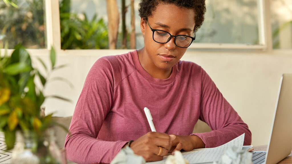 Pessoa estudando em casa, escrevendo anotações no caderno para se qualificar para o mercado de trabalho, com notebook ao lado.