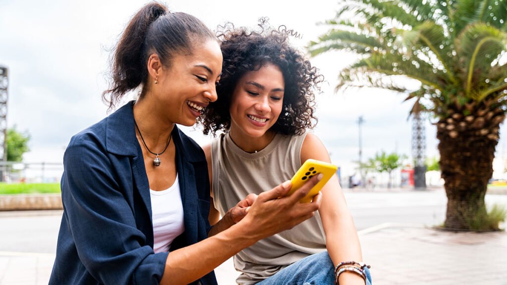 Duas mulheres sorrindo enquanto olham juntas para um celular amarelo, representando a conexão genuína com o público por meio do marketing emocional.