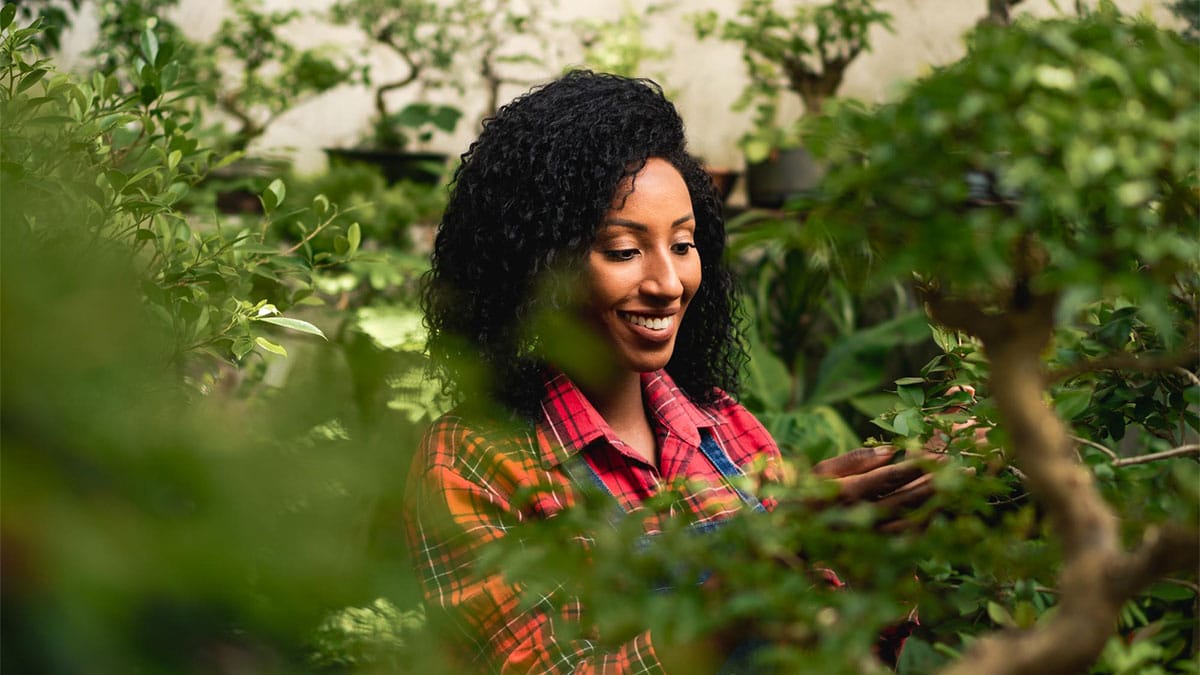 Mulher sorridente cuidando de plantas em meio ao verde, representando o conceito de empreendedorismo verde e a conexão entre sustentabilidade e trabalho feminino.