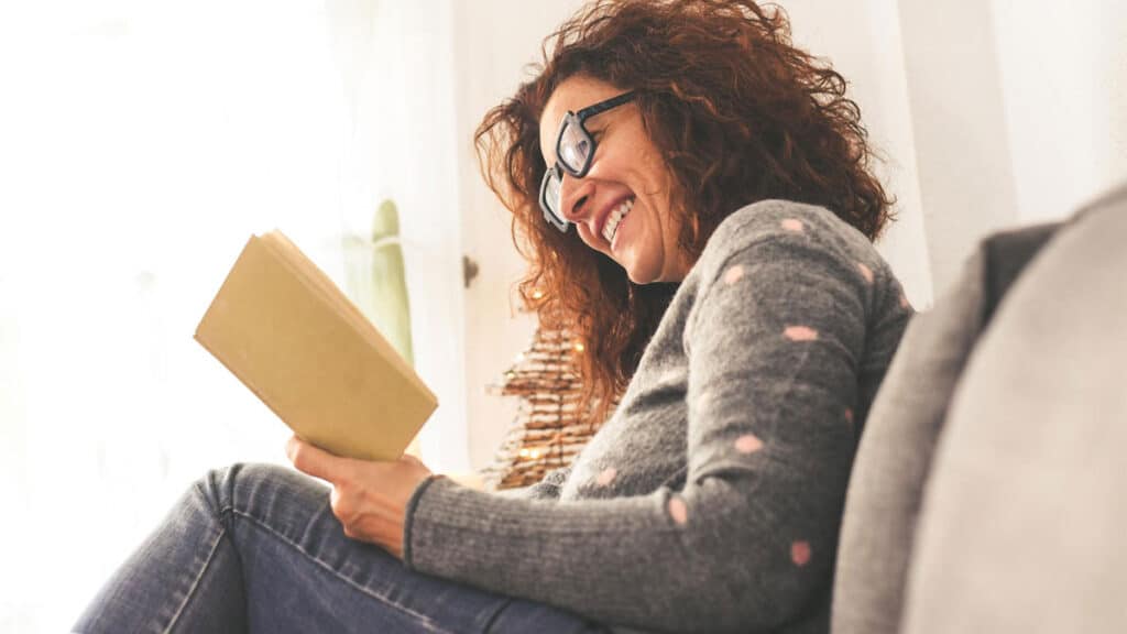Mulher sorridente lendo um livro em casa, representando o poder da leitura e o incentivo aos livros para mulheres empreendedoras.