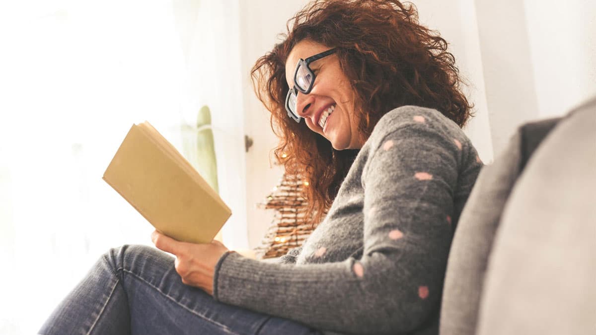 Mulher sorridente lendo um livro em casa, representando o poder da leitura e o incentivo aos livros para mulheres empreendedoras.