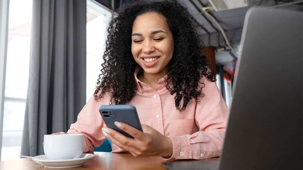 Mulher sorridente segura o celular enquanto trabalha em um café, simbolizando o uso de tendências no marketing digital para impulsionar negócios online.